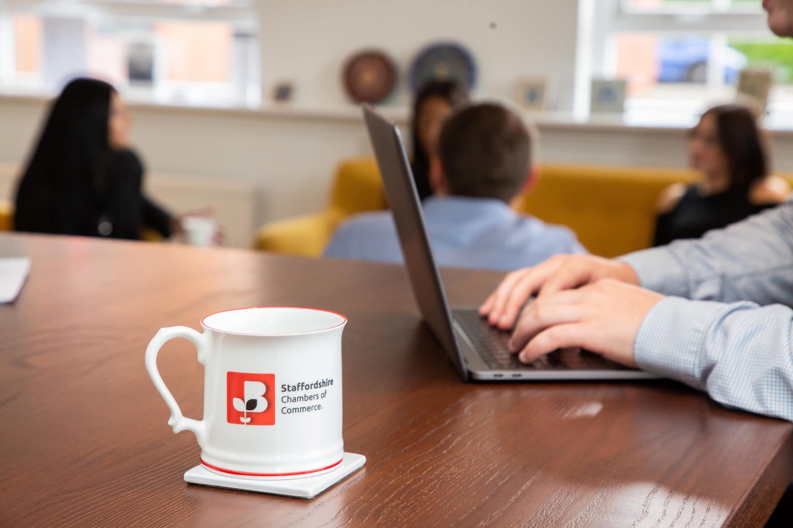 Staffordshire Chambers' Logo on a mug on a table next to a man working on his laptop