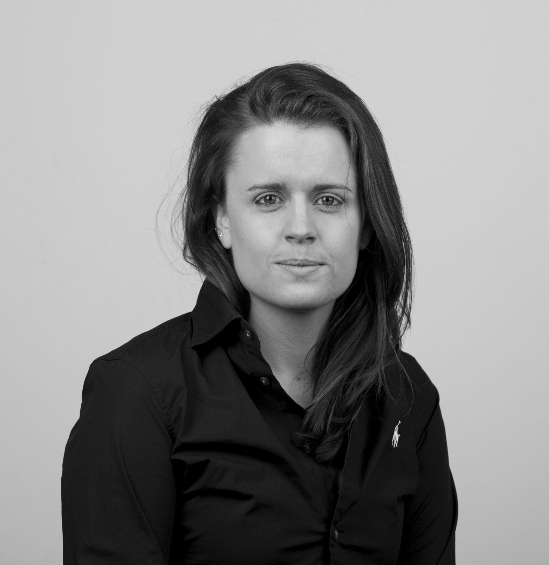 Black and white portrait of Josie Morris, with long hair, wearing a dark collared shirt with a small logo, looking directly at the camera with a neutral expression.