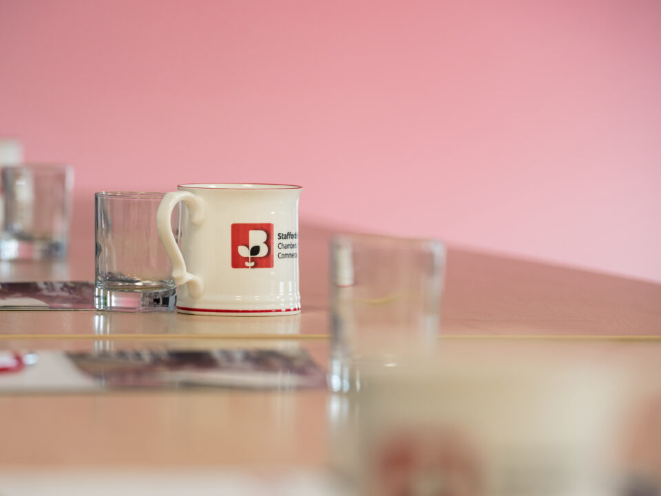 Close-up of a Staffordshire Chambers of Commerce branded mug on a table, with a pink background and glass cups nearby.