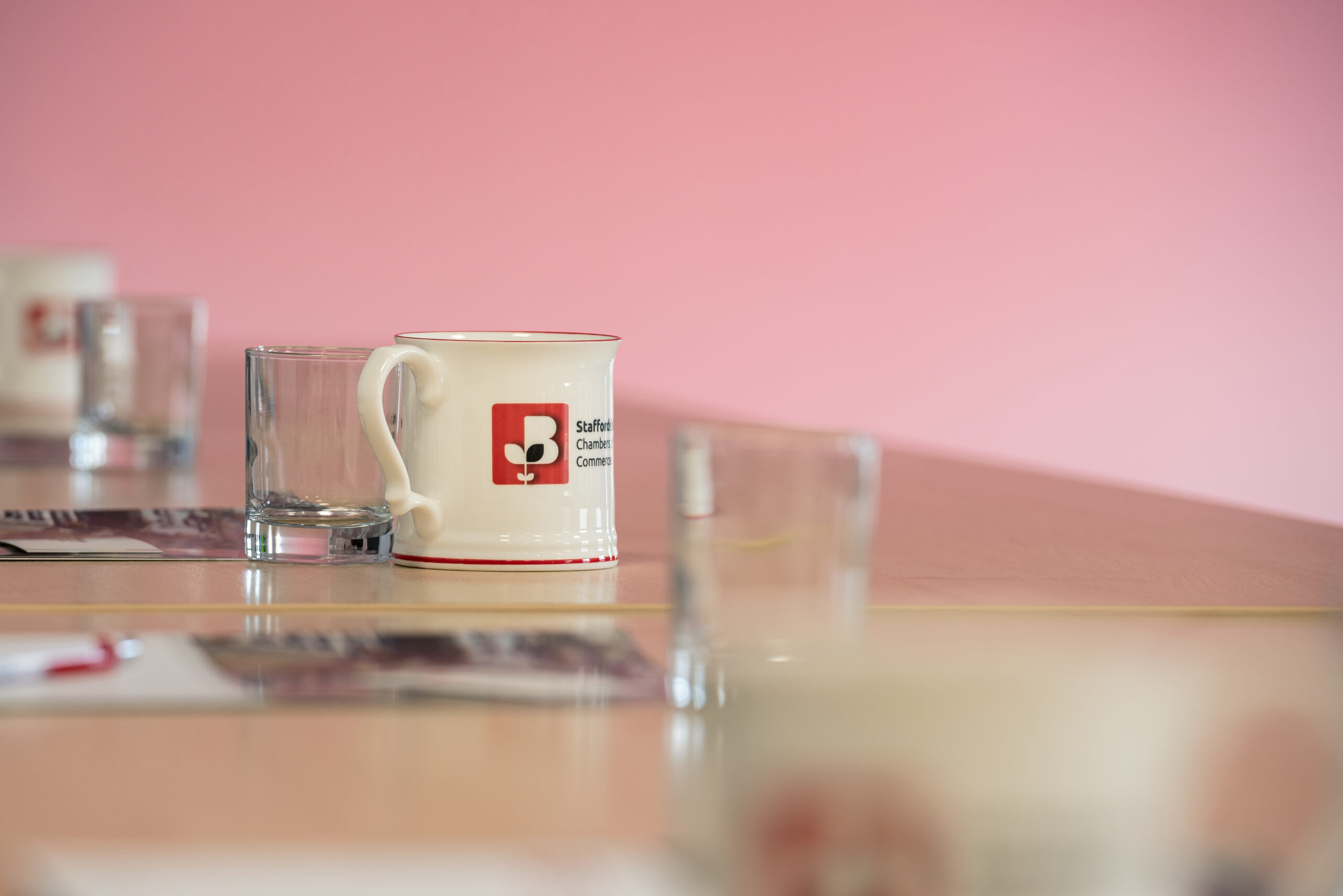 Close-up of a Staffordshire Chambers of Commerce branded mug on a table, with a pink background and glass cups nearby.