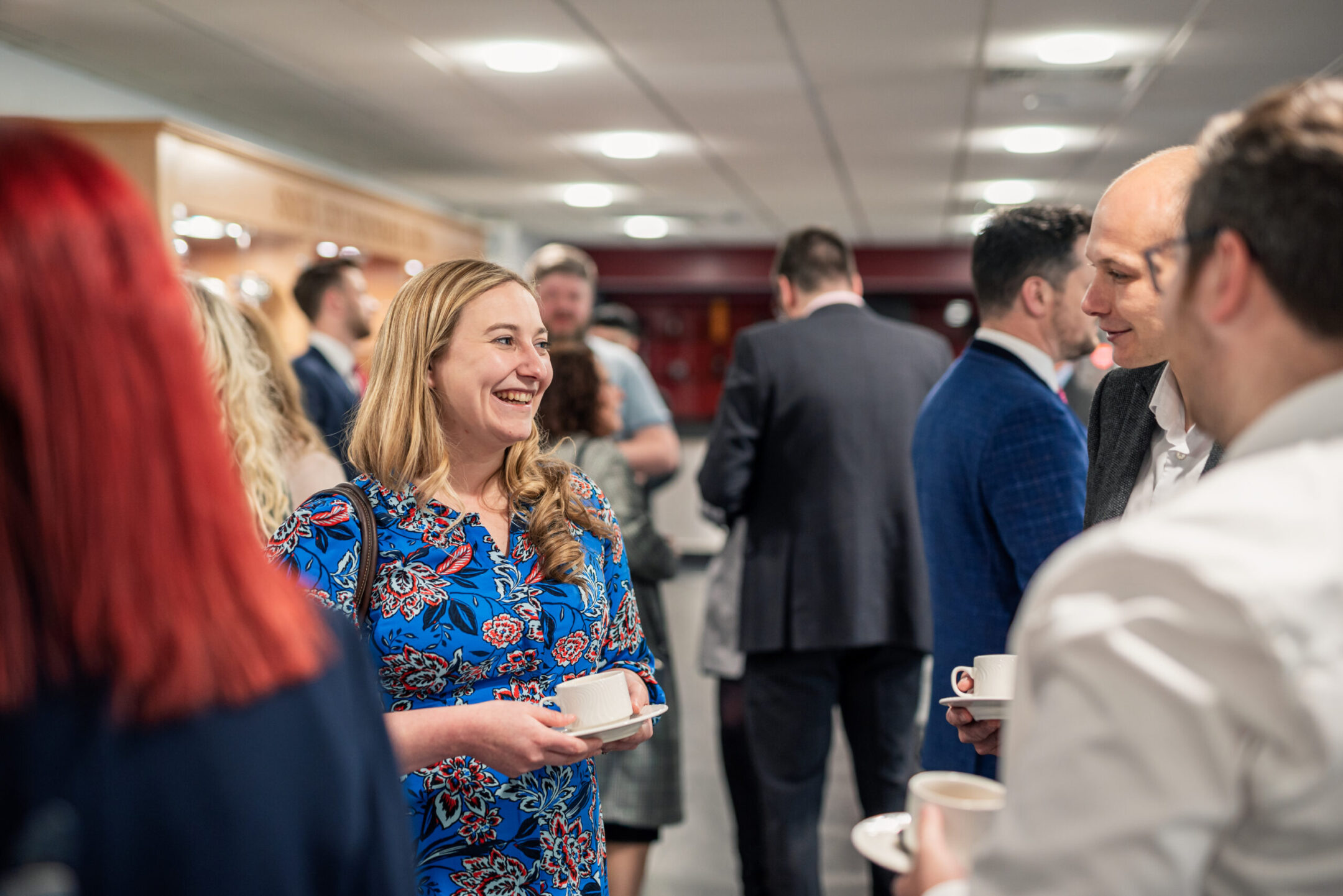A smiling woman in a blue floral dress holding a cup and saucer while networking at a Staffordshire Chambers of Commerce event, with attendees in formal attire conversing in the background.