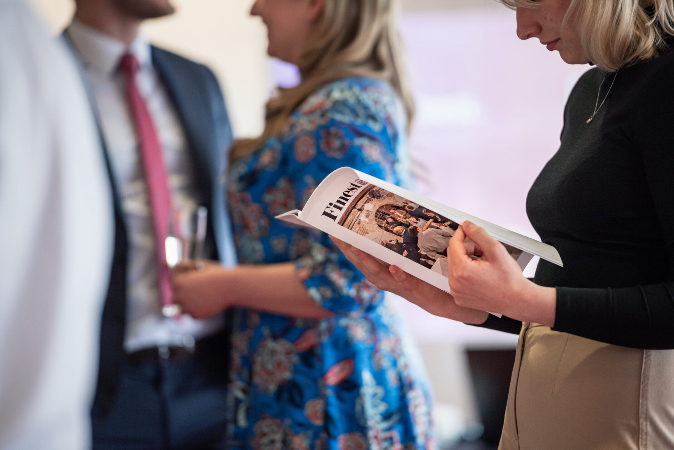 A person in a black top and beige trousers holding and reading a printed magazine at a Staffordshire Chambers of Commerce event, with attendees conversing in the background.