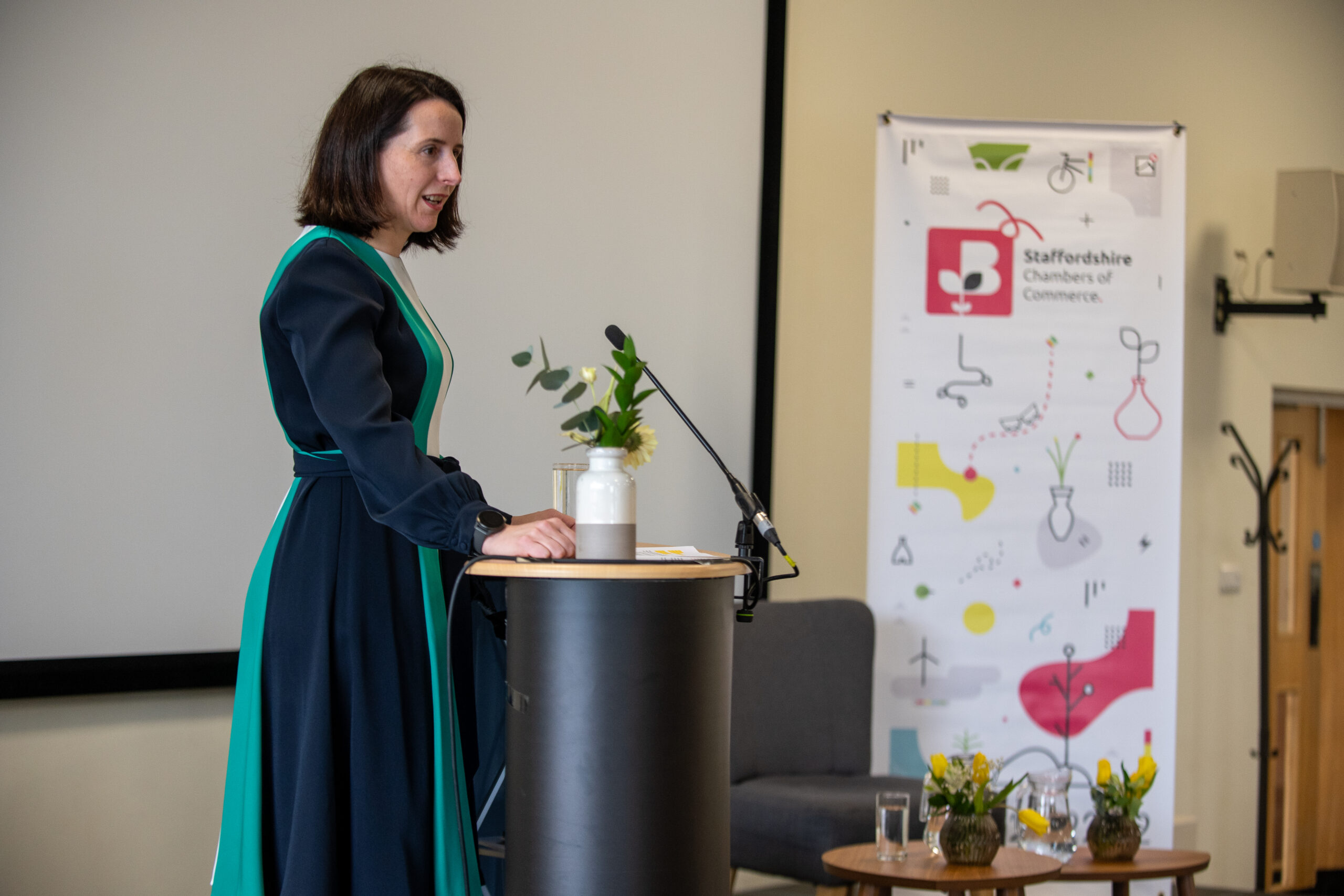 Rachel Laver, CEO of Staffordshire Chambers of Commerce, presenting at a podium during a Chamber event, with a branded banner and decorative flowers in the background.