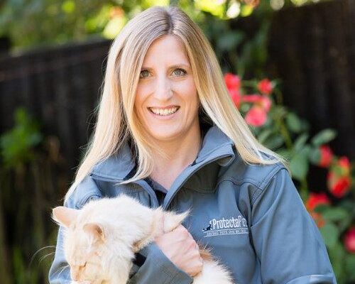 Eve Davies smiling outdoors, holding a fluffy cream-colored cat while wearing a grey jacket, with a garden and red flowers in the background.