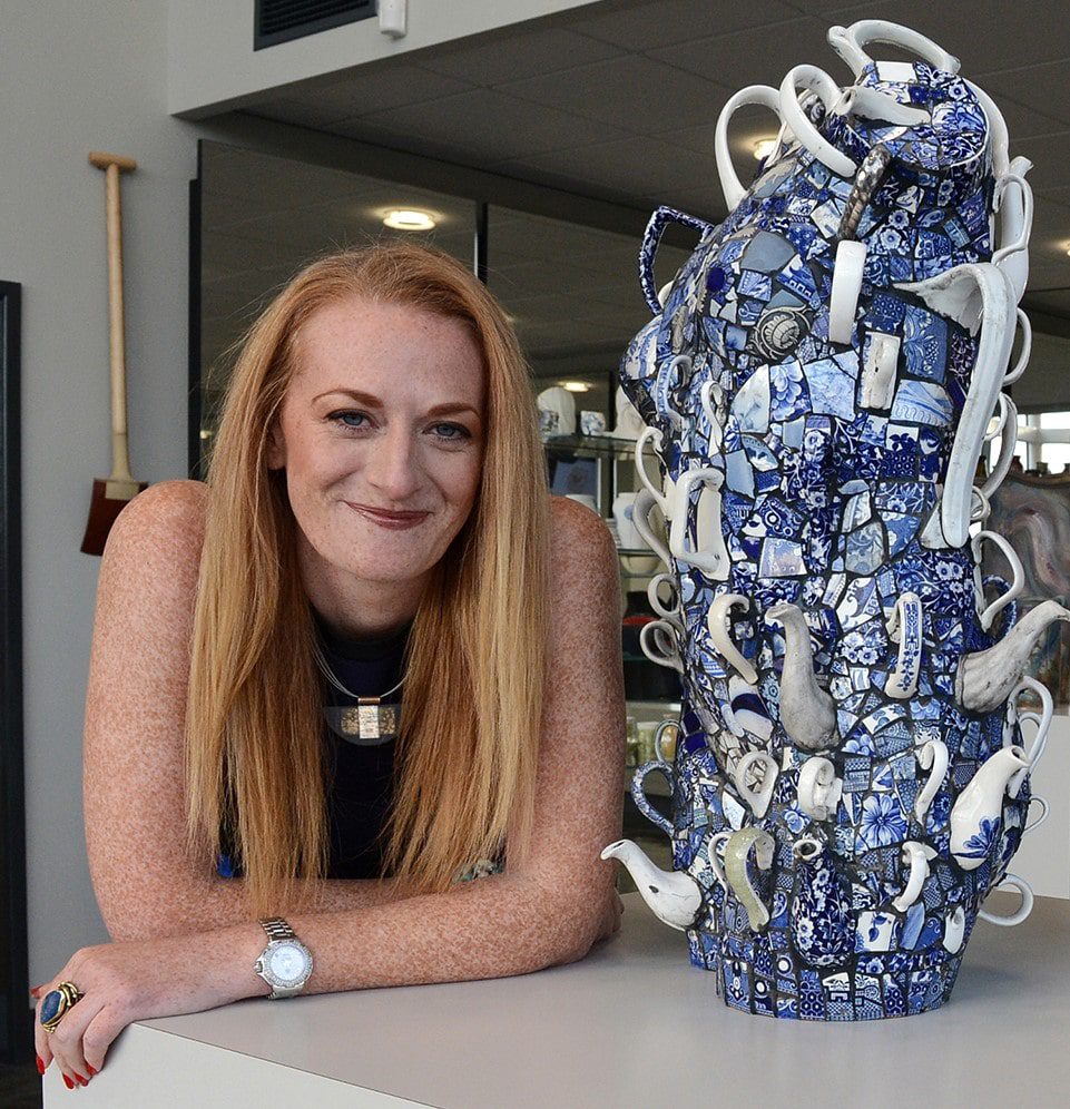 Hannah Ault smiling at the camera, leaning on a table beside an intricate blue and white ceramic sculpture made from mosaic and porcelain pieces.