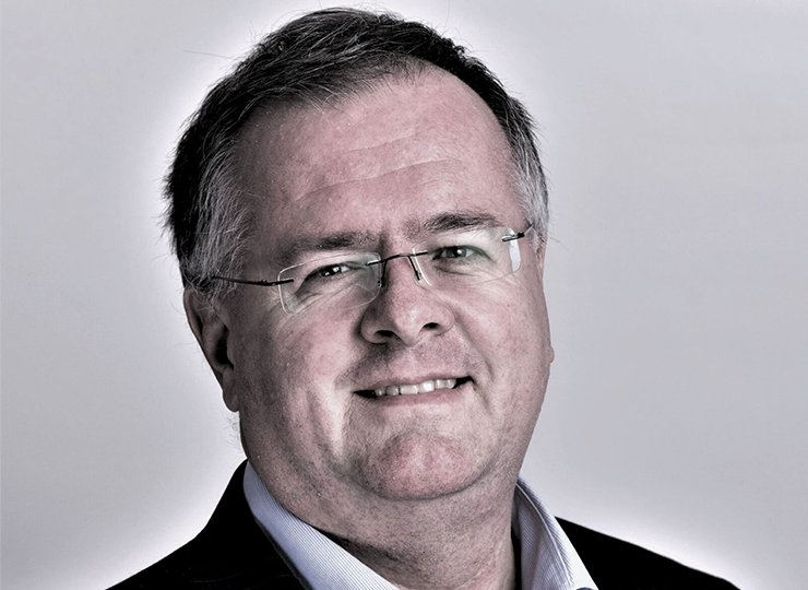 Lee White, smiling at the camera, wearing glasses, a dark pinstripe suit, and a light-colored shirt, against a neutral grey background.