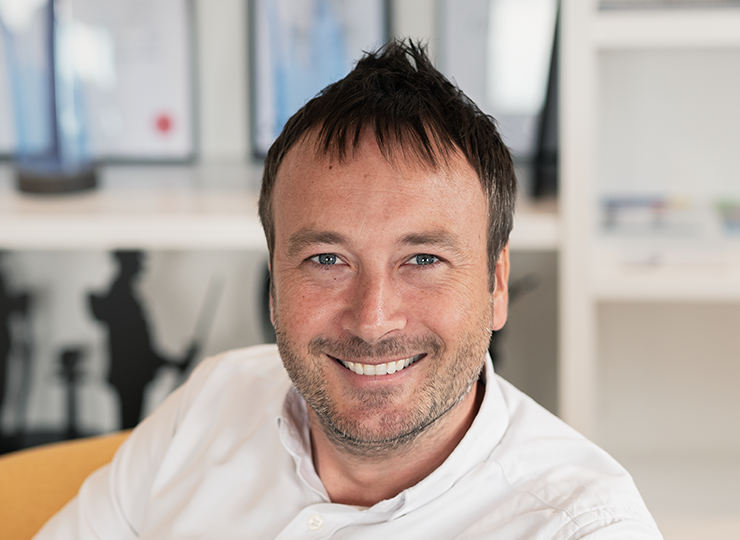 Matt Hubbard, smiling at the camera, with short dark hair, wearing a white shirt, in a modern office setting with shelves and framed certificates in the background