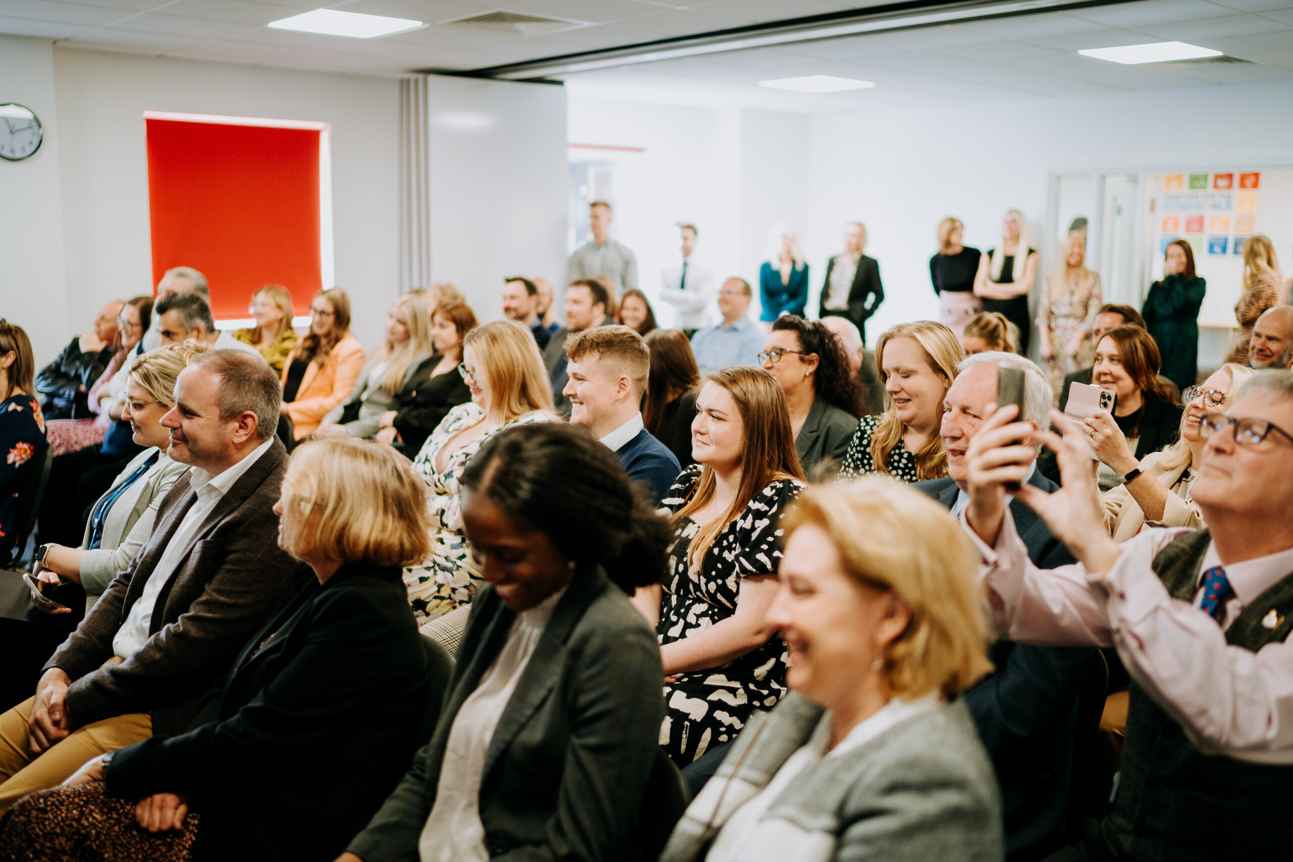 Attendees at a Staffordshire Chambers of Commerce event seated in a room, smiling, listening, and engaging, with some capturing moments on their phones.