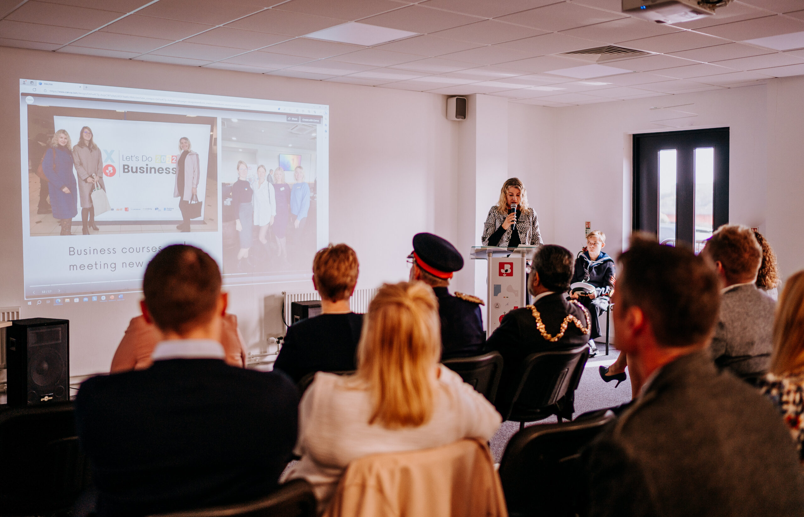 A business event at Staffordshire Chambers of Commerce, showing a speaker presenting at a podium while an audience listens, with a large screen displaying a group photo and event details.