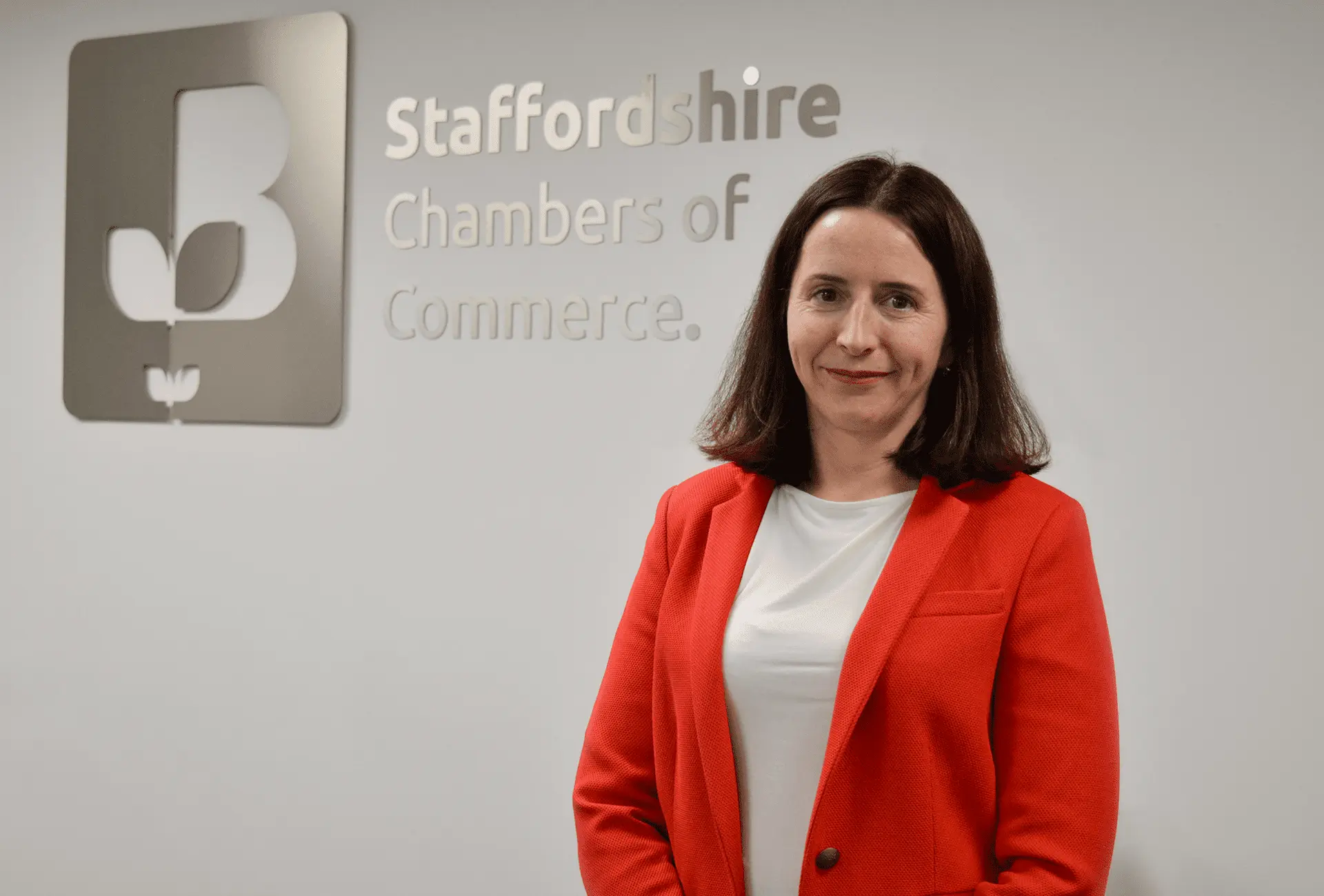 Rachel Laver, CEO of Staffordshire Chambers of Commerce, standing in front of the Staffordshire Chambers logo, wearing a red blazer and white blouse, smiling at the camera.