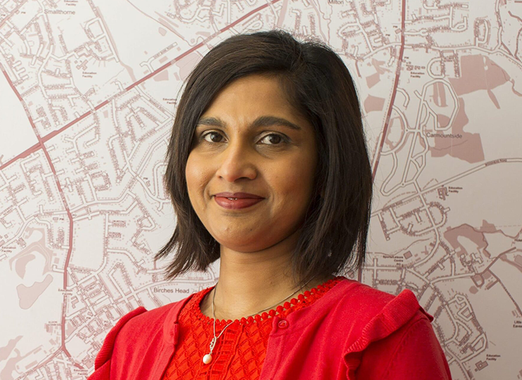 Ramona Hirschi, with shoulder-length dark hair, wearing a red cardigan and dress, standing in front of a detailed map background.