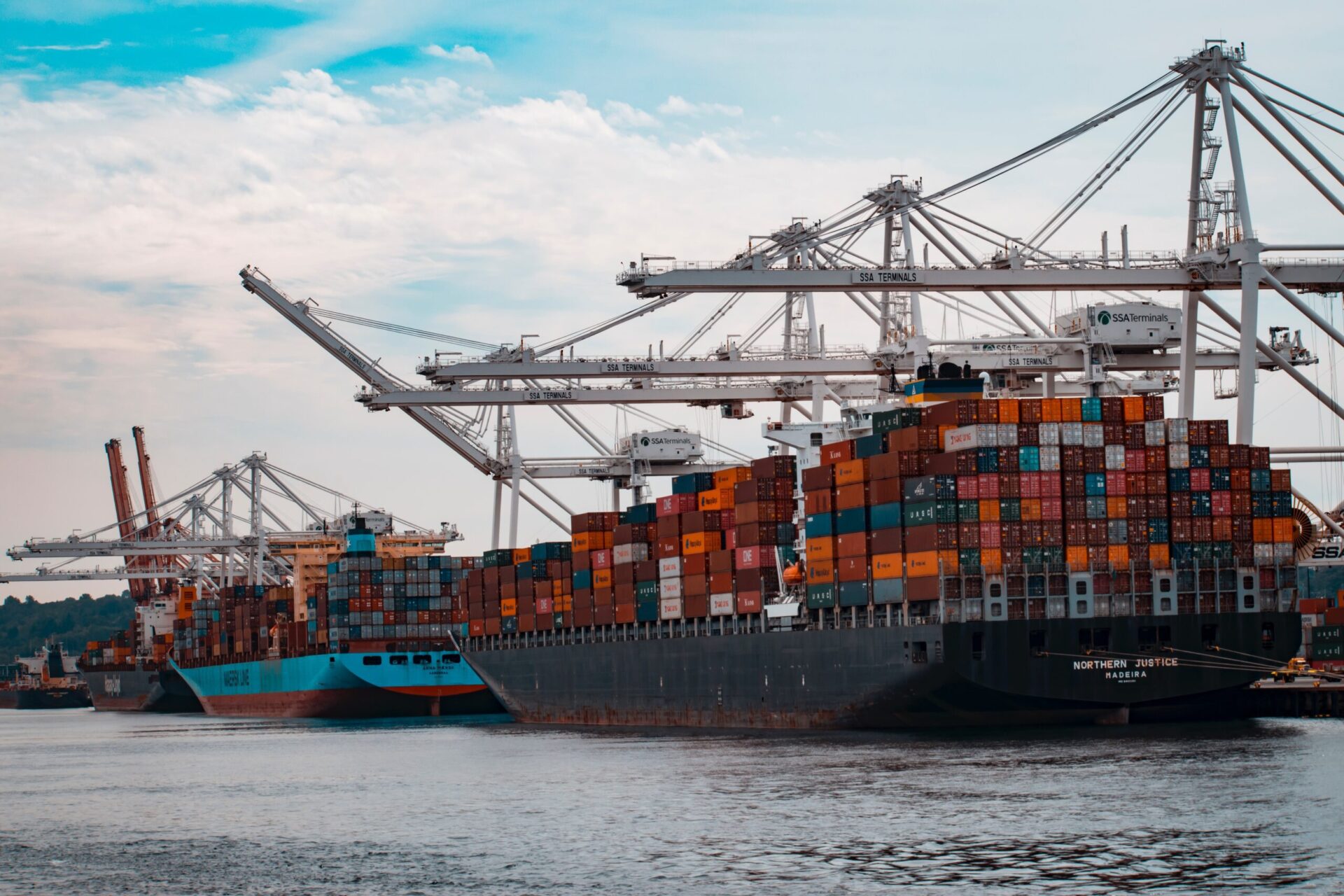 Cargo ships loaded with colorful shipping containers docked at a port with large white cranes and SSA Terminals signage.