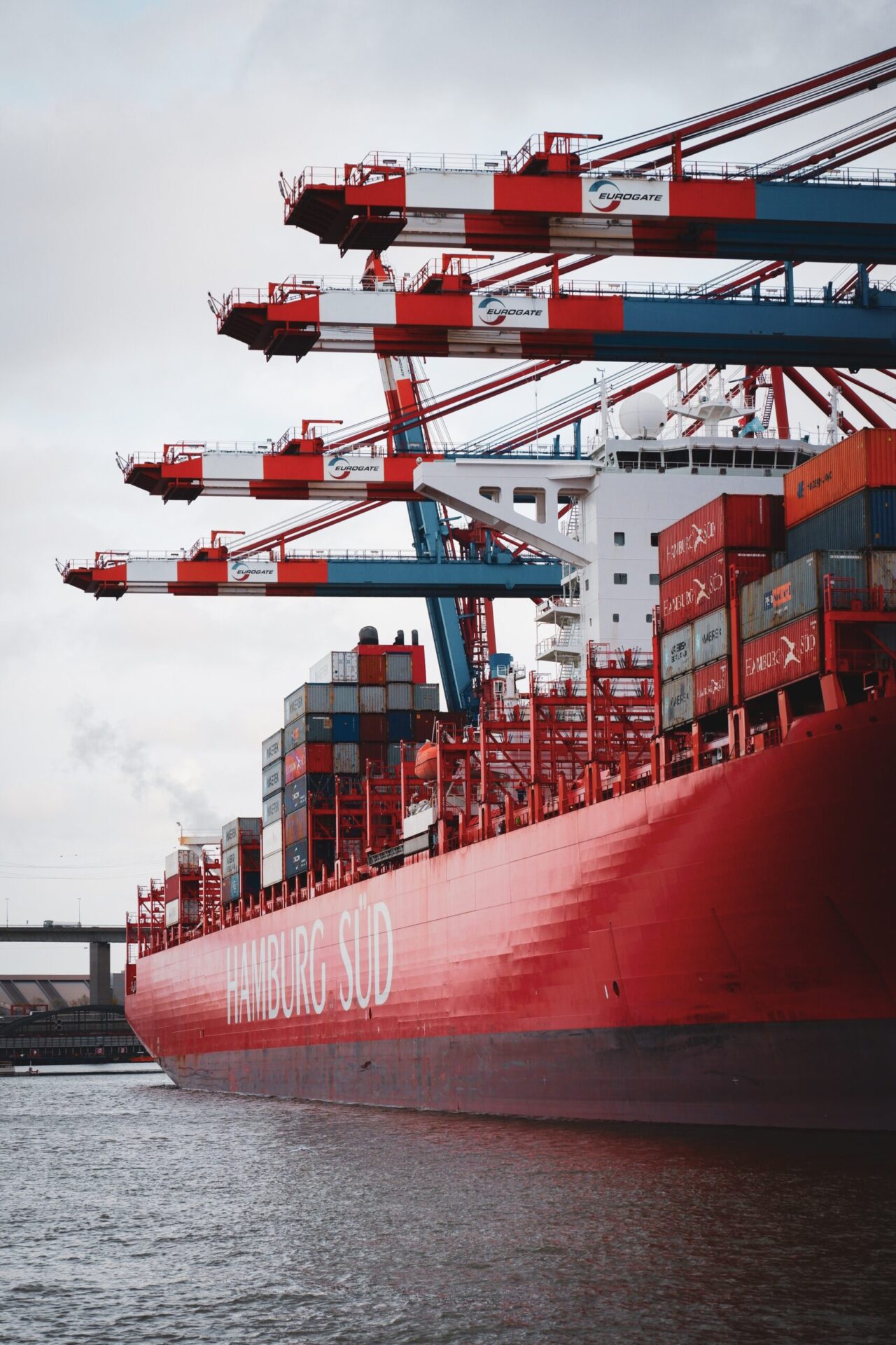 Large red cargo ship labeled 'Hamburg Süd' with stacked shipping containers and red-and-blue cranes at a port.