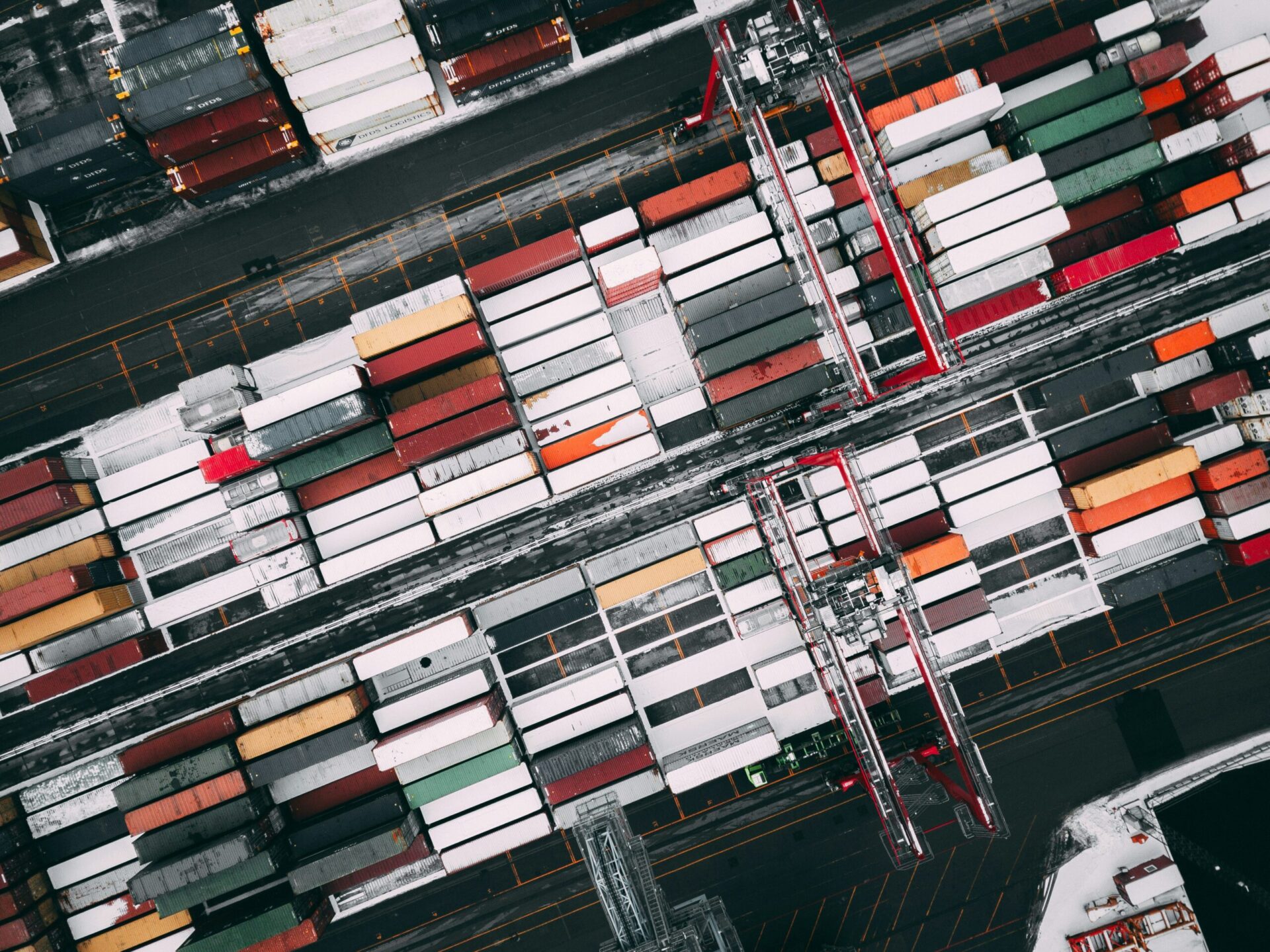 Aerial view of a shipping yard with rows of colorful cargo containers and red cranes on a snowy day.