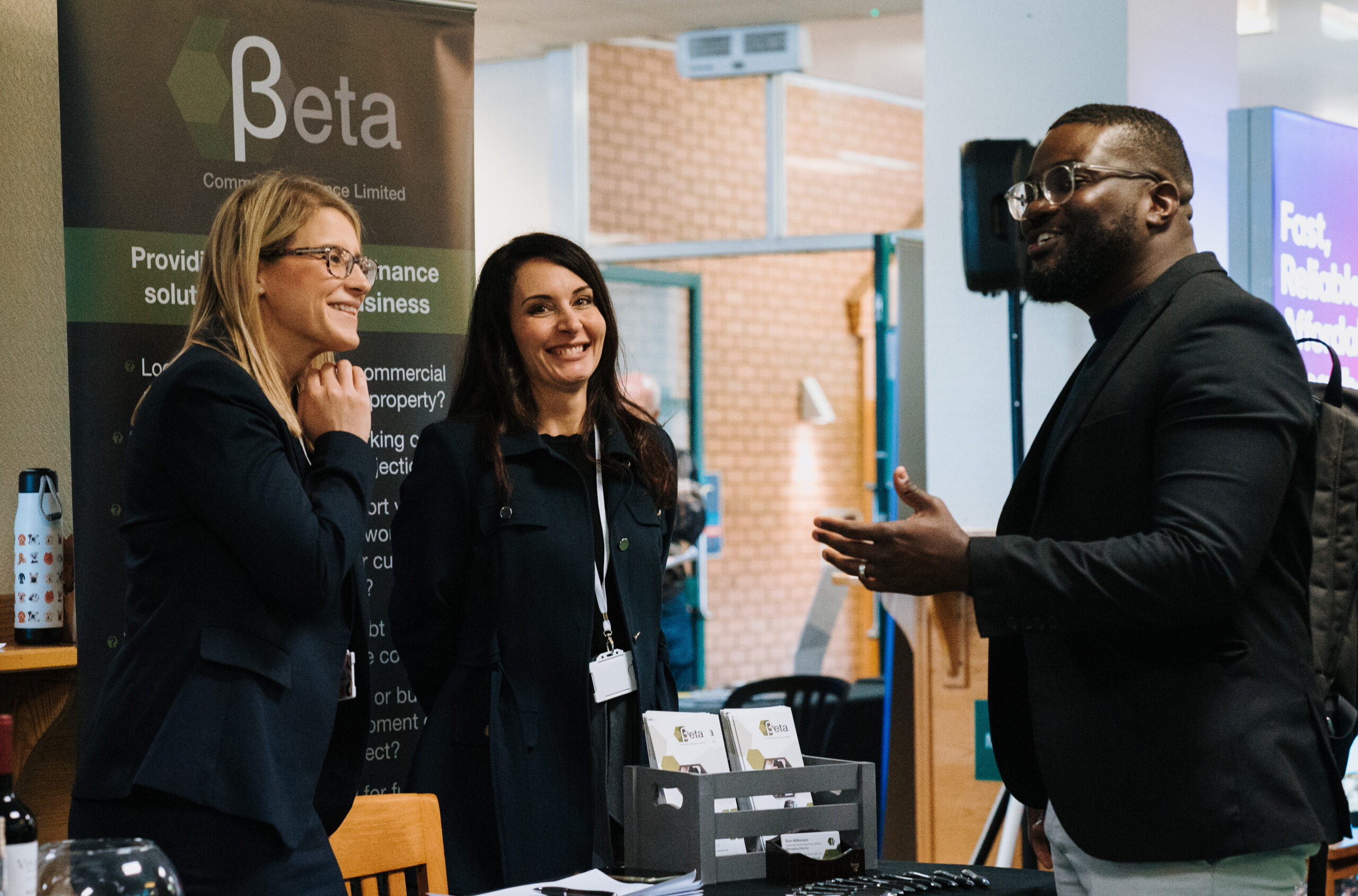 A professional networking discussion at a Staffordshire Chambers of Commerce event, featuring two women at a booth for Beta Commercial Finance Limited and a man engaging in conversation.