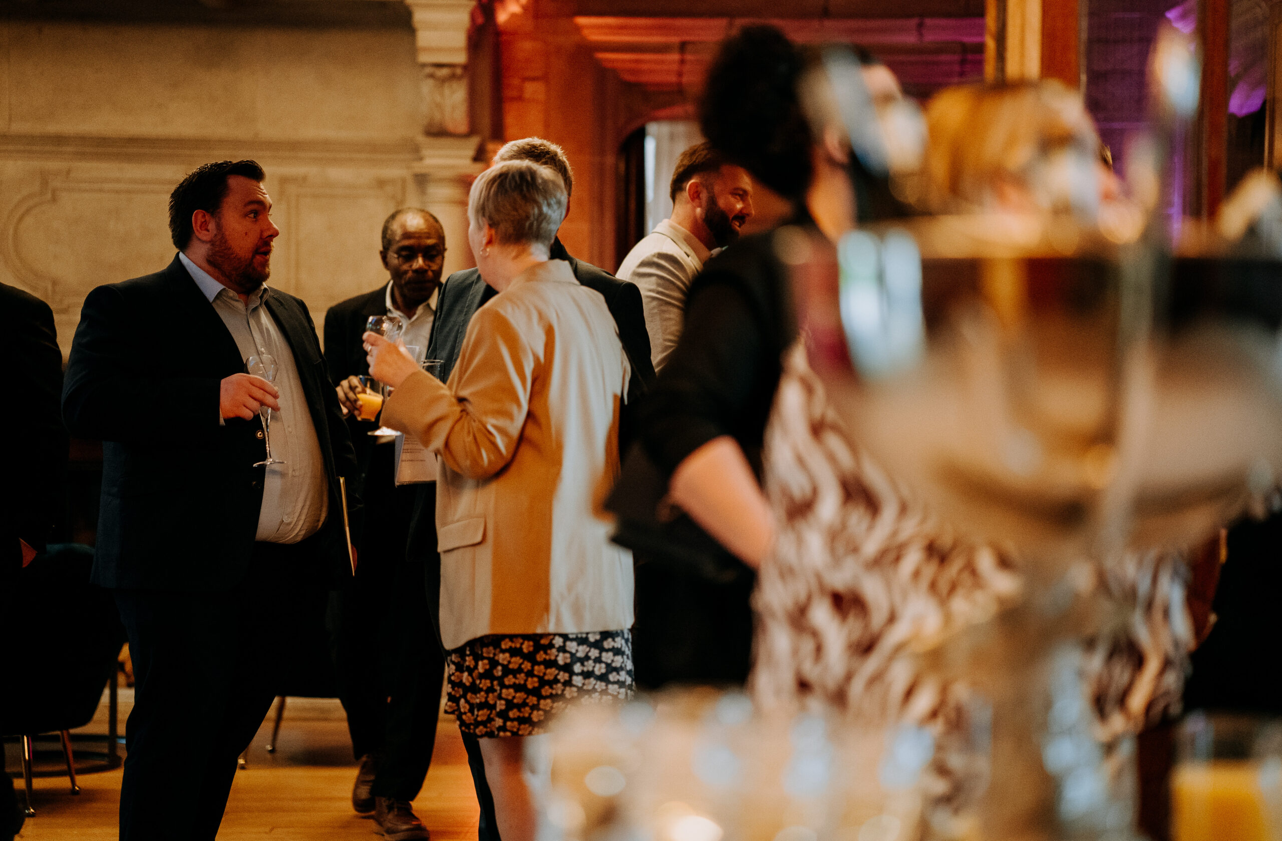 A group of individuals networking during a social event, holding glasses of beverages and engaging in conversations in a warmly lit room.