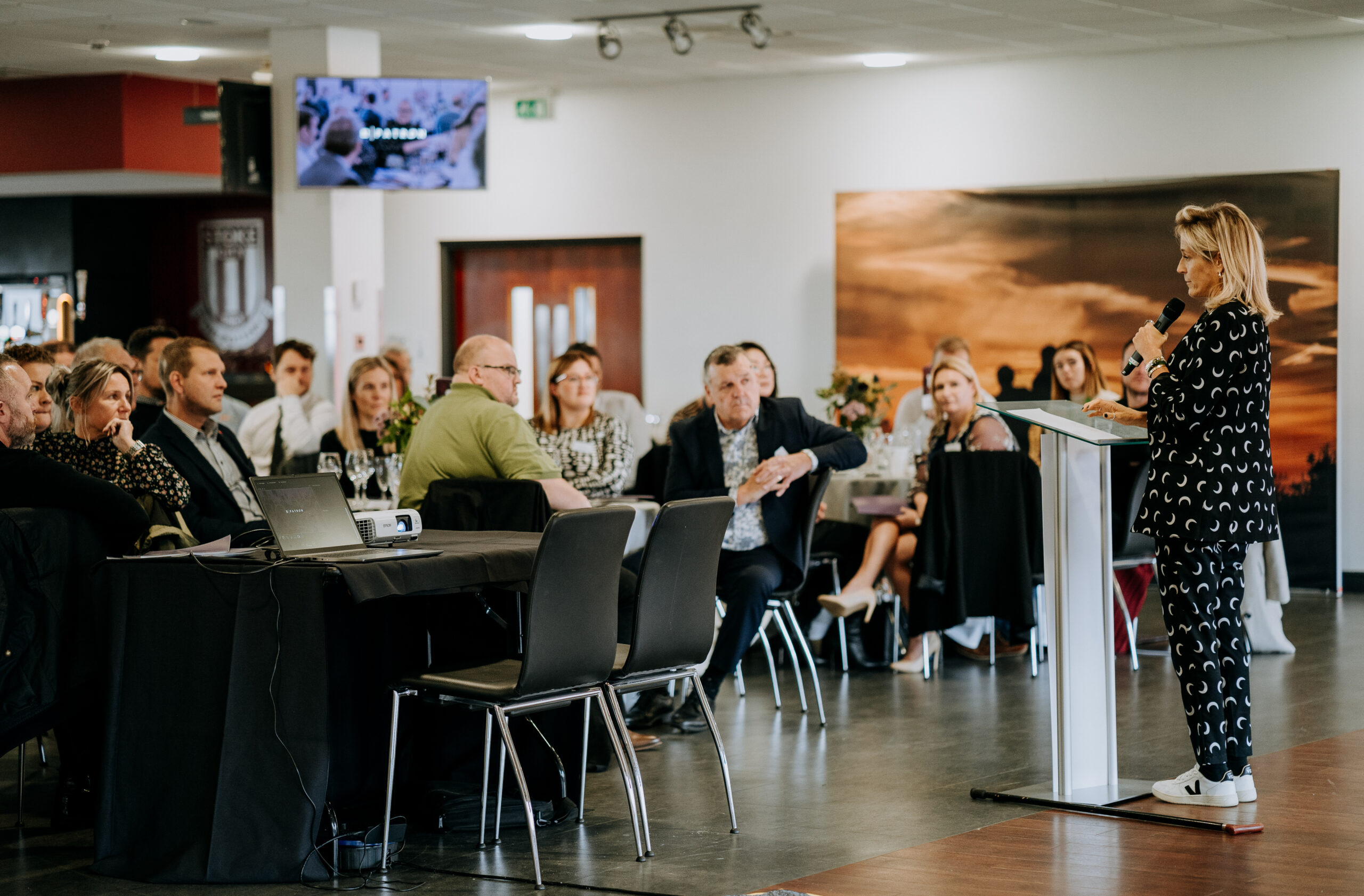 Speaker addressing the audience at a Staffordshire Chambers of Commerce event, with attendees seated at round tables, a projector on a table, and a large wall artwork in the background.